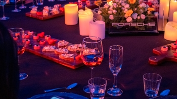Primer plano de una mesa de catering en un evento Porsche: tabla de canapés, copas de vino, vasos y velas, con un arreglo floral que muestra el logo Porsche iluminado.