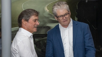 Dos hombres conversando en el evento de inauguración, con un fondo digital de una pista de carreras, sugiriendo discusiones sobre automovilismo.