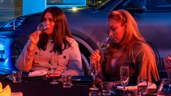 Dos mujeres degustan vino en una mesa frente a un Porsche durante un evento; se observan copas, vasos y la carrocería del vehículo iluminada por luces de colores.