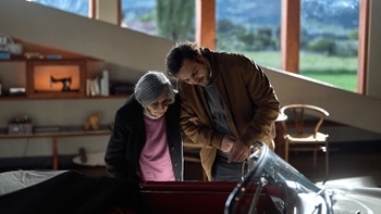 Un hombre joven con barba y una mujer mayor con canas observan con interés el interior de un Porsche 356 convertible clásico. Ambos están inclinados sobre el parabrisas, mirando el habitáculo que parece tener tapicería roja. Se encuentran dentro de un espacio bien iluminado con grandes ventanales que muestran un paisaje verde al fondo.