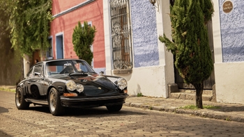 Un Porsche 911 clásico de color negro se encuentra estacionado en una calle adoquinada, flanqueado por edificios de colores vivos: uno rosa o terracota y otro azul claro. Hay árboles pequeños y vegetación junto a las fachadas. La escena irradia un ambiente pintoresco de un pueblo o ciudad antigua.
