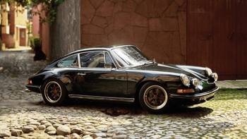 Un Porsche 911 clásico de color negro brillante está estacionado sobre una calle empedrada. El coche presenta detalles cromados, faros redondos distintivos y llantas de color bronce. Al fondo, se aprecia una pared de piedra rojiza y vegetación, sugiriendo un entorno histórico o rústico.