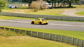 Porsche 718 Cayman GT4 amarillo en una pista de carreras, con vista a la ambulancia en el fondo.