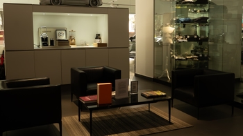 Un área de espera en un concesionario Porsche, equipada con tres sillones individuales de cuero negro y una mesa de centro con libros y folletos. Al fondo, se aprecia una estación de café Nespresso y una vitrina de cristal con merchandising de la marca y un maniquí.