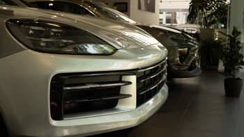 Vista frontal parcial de dos Porsche Cayenne alineados en un concesionario. En primer plano, un Porsche Cayenne de color plateado o gris claro, mostrando sus faros y parrilla. Detrás, se vislumbra otro Porsche Cayenne oscuro. Una planta decorativa se encuentra a la derecha.