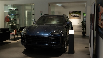 Un Porsche Cayenne de color azul marino metálico, estacionado majestuosamente en el centro de un showroom o concesionario. A la izquierda, se observa una vitrina con merchandising de la marca. A la derecha, una planta decorativa y un cuadro con la imagen de un Porsche clásico.