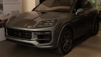 Detalle frontal de un Porsche Cayenne Coupé de color gris oscuro exhibido en un concesionario. Se aprecian sus faros, la parrilla frontal y parte de la rueda delantera. Al fondo, un cartel promocional anuncia "EL NUEVO CAYENNE - EMOCION GARANTIZADA".