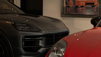Vista en primer plano de dos vehículos exhibidos en un concesionario. En el lado izquierdo, un Porsche Cayenne de color gris oscuro metálico. A la derecha, parcialmente visible, se asoma la parte frontal de un deportivo rojo. Al fondo, se aprecia una pintura de un coche de carreras naranja.