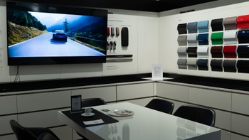 Interior de una sala de configuración en un concesionario Porsche, con una pantalla mostrando un Porsche deportivo azul en una carretera montañosa. En primer plano, una mesa de diseño moderno con un café y dulces, rodeada de sillas negras. A la derecha, una pared exhibe muestras de pintura y materiales, mientras que a la izquierda se ven accesorios de vehículos.