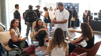 Grupo de mujeres conversando en el showroom de Porsche, revisando documentos y casco en mano, con asistentes y coches al fondo durante un evento.