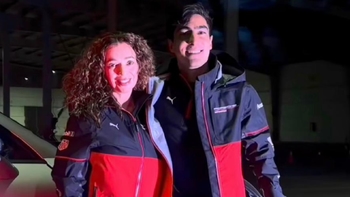 Un hombre y una mujer sonrientes posan juntos, ambos vistiendo chaquetas con logotipos de Porsche y otros patrocinadores. La imagen está tomada en un entorno oscuro, posiblemente de noche o en un interior con poca luz.
