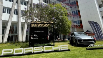 Un Porsche Macan plateado se exhibe al aire libre sobre un césped verde, frente a edificios modernos. Un gran letrero con el nombre "PORSCHE" se encuentra en el pasto, junto a una carpa de exhibición negra y banderas con los logos de Porsche. Un árbol de jacarandá con flores moradas es visible en el fondo.
