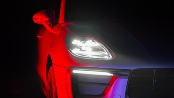 Primer plano de la parte frontal de un Porsche Macan, destacando el faro delantero encendido y las luces diurnas LED. El coche está iluminado por luces ambientales de colores rojo y púrpura que realzan sus contornos en la oscuridad.