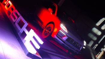 Un Porsche Macan se exhibe en un ambiente interior con iluminación dramática en tonos rojos y púrpuras. Un gran letrero iluminado con el nombre "PORSCHE" se encuentra en primer plano, inclinado. Los faros del coche están encendidos, resaltando su diseño frontal.