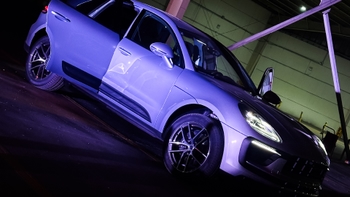 Un Porsche Macan se exhibe en un ambiente interior oscuro bajo iluminación ambiental de color púrpura. Ambas puertas delanteras del coche están abiertas, y sus faros están encendidos, resaltando su presencia.