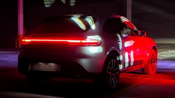 Un Porsche Macan gris oscuro se muestra desde la parte trasera en un estacionamiento subterráneo. Las luces traseras LED rojas brillan intensamente, y el coche está bañado en proyecciones de luz rojas y patrones de luz azules y blancas, creando un efecto dramático y moderno.