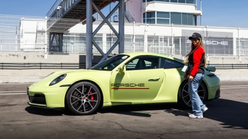 Porsche 911 color lima estacionado en el pit lane con la puerta abierta y una mujer con gorra y chaqueta roja junto al coche.