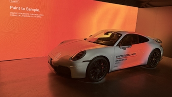 Porsche 911 GTS blanco expuesto frente a una pantalla naranja con el texto "Paint to Sample", iluminación ambiental en un espacio interior.