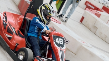 Niño con casco y camiseta azul al volante de un go-kart rojo K1 Speed con el número 48, conduciendo en una pista.