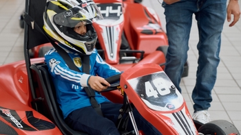 Niño con casco y camiseta azul sentado al volante de un go-kart rojo K1 Speed con el número 48, con un adulto a su lado.