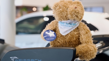Oso de peluche Make-A-Wish México con pañuelo y sticker, sobre un spoiler Weissach, con un Porsche blanco de fondo borroso.