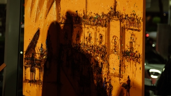 Detalle de una pintura naranja con textura y salpicaduras; sombra proyectada de espectadores sobre la superficie y reflejo de luces del showroom.