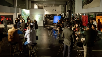 Panorámica del evento en showroom/galería: asistentes en mesas altas, obras colgadas y un ambiente nocturno con iluminación cálida; vehículo cubierto al fondo.
