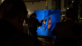 Performance de pintura en vivo sobre lienzo azul en showroom; espectadores en silueta y pinceladas naranjas.
