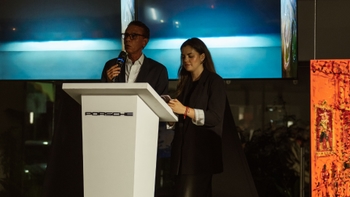 Dos presentadores en un podio con el logo "Porsche", pantalla digital y obras de arte al fondo durante un evento en showroom; personas no identificadas.