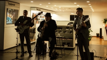 Trío musical en vivo (saxofón, bajo eléctrico y guitarra) actuando en un showroom; iluminación tenue, micrófonos y cables en el suelo, público fuera de plano.