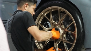 Un técnico con guantes naranjas limpia meticulosamente la rueda dorada de un vehículo Porsche en un taller especializado.