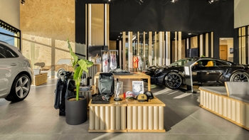 Vista interior de un moderno showroom de Porsche Now, con un Porsche 911 negro en exhibición sobre una plataforma, un Porsche Cayenne blanco parcialmente visible a la izquierda, y una mesa central con merchandising de la marca Porsche.