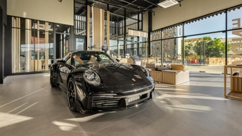 Un Porsche 911 negro brillante estacionado prominentemente en el centro de un luminoso y moderno showroom de Porsche, con grandes ventanales que permiten la entrada de luz natural.
