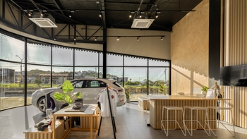 Interior amplio y luminoso de un showroom de Porsche, mostrando un Porsche Cayenne de color claro cerca de los grandes ventanales, un área de atención con barra de madera y taburetes, y elementos de merchandising de la marca.