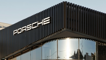 Primer plano angular del exterior de un moderno concesionario Porsche, destacando su fachada de listones oscuros y una esquina curvada de cristal, con el logotipo "PORSCHE" en letras blancas.