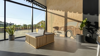 Interior de una zona de descanso o sala de espera en un showroom de Porsche, con grandes ventanales que ofrecen una vista exterior, un sofá moderno y un llamativo mural de diseño floral con la silueta de un coche clásico en la pared.