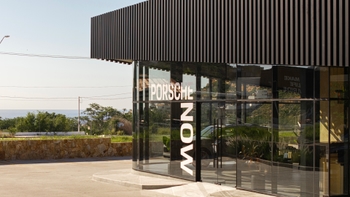 Vista exterior de un moderno showroom "Porsche Now" con su distintiva fachada de listones oscuros y ventanales de cristal que muestran el logotipo de la marca, con vistas al mar y la carretera de fondo.