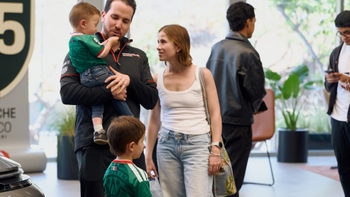 Un hombre sostiene a un bebé vestido con un jersey verde de la selección mexicana, mientras una mujer y otro niño con un jersey similar se encuentran cerca, en lo que parece ser un evento o concesionario de Porsche, con automóviles y otras personas de fondo.