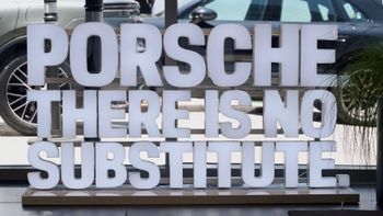 Un letrero luminoso de gran tamaño con las palabras "PORSCHE THERE IS NO SUBSTITUTE" en letras blancas, visible en un concesionario o sala de exposición, con automóviles Porsche borrosos al fondo a través de un cristal y una planta a la derecha.
