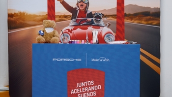Mesa Make-A-Wish y Porsche con el lema "Juntos Acelerando Sueños". Sobre ella, artículos de manualidades (pinturas, pinceles), un osito de peluche y coches de juguete.