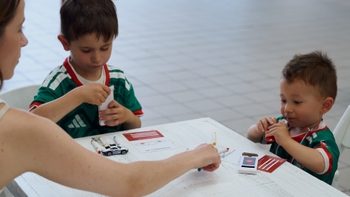 Una mujer, vista de espaldas, comparte una mesa blanca con dos niños pequeños. El niño mayor, a la izquierda, viste una camiseta verde y blanca y parece estar concentrado en una actividad manual. El niño más joven, a la derecha, también con una camiseta verde y blanca, mira atentamente mientras sujeta algo pequeño. Sobre la mesa hay rotuladores y un pequeño coche de juguete.