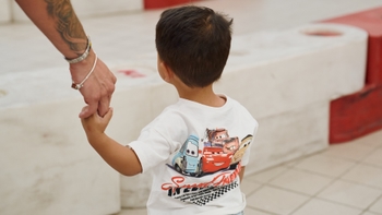 Un niño pequeño, visto de espaldas, usa una camiseta blanca con personajes de "Cars" y sostiene la mano de un adulto cuyo brazo tatuado es visible. El fondo muestra una zona con barreras rojas y blancas, sugiriendo un área de juego o pista.