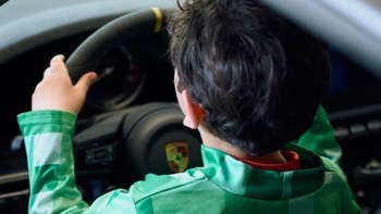 Niño con traje verde de espaldas, manos al volante de un Porsche, mostrando el emblemático escudo y un detalle amarillo.