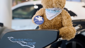 Oso de peluche marrón con bandana y sticker de 'Make-A-Wish México', sentado sobre el alerón trasero de un vehículo Porsche con el distintivo 'Weissach RS'.