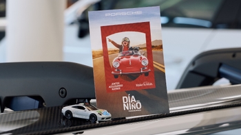 Una tarjeta promocional vertical de Porsche, celebrando el "Día del Niño" y la colaboración con "Make A Wish", descansa sobre una superficie de fibra de carbono. La tarjeta muestra a un niño con gafas de aviador sonriendo desde un Porsche descapotable antiguo. Delante de la tarjeta, un pequeño coche de juguete blanco de rally, similar a un Porsche, se apoya en la misma superficie.