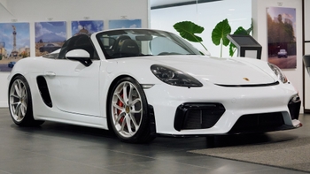 Un Porsche Spyder convertible blanco, con llantas plateadas y pinzas de freno rojas, se exhibe en un luminoso concesionario. El fondo muestra impresiones en la pared de paisajes urbanos y plantas verdes, creando un ambiente de concesionario sofisticado.