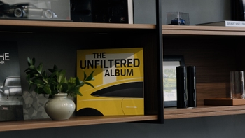 Estantería decorativa con un libro "The Unfiltered Album", una planta en maceta y otros objetos, en un ambiente relacionado con Porsche.