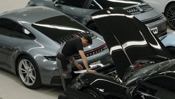 Mecánico revisando el motor de un Porsche negro con el capó abierto, junto a un Porsche 911 Carrera gris en un taller de servicio.