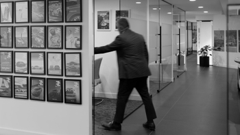 Pasillo de oficina con hombre en movimiento y una pared decorada con una serie de pósteres enmarcados de la historia y modelos Porsche.