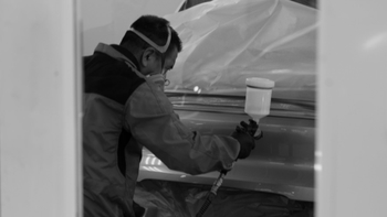 Pintor automotriz Porsche aplicando capa de pintura a una pieza de coche cubierta con plástico en un taller de carrocería.